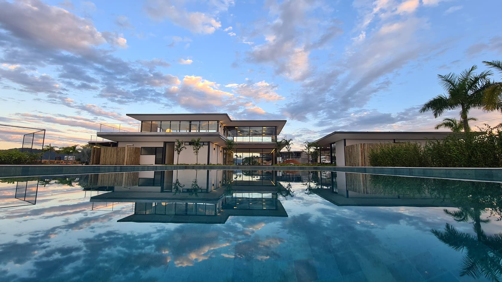 Edifício do clube Reserva das Aroeiras, de vidro e concreto, refletido na piscina ao entardecer.